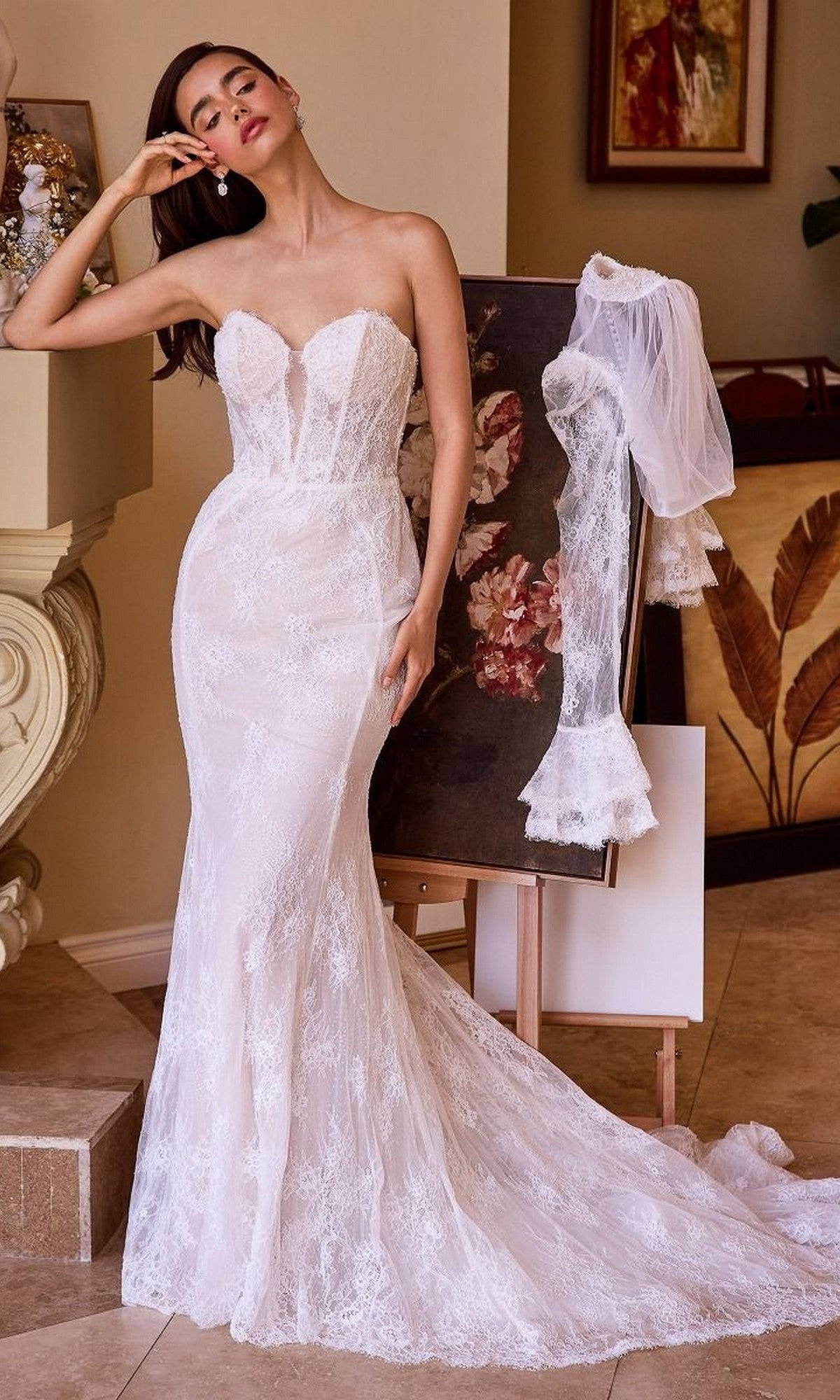 A woman in the Andrea and Leo WL042 long wedding dress with a strapless lace fit-and-flare silhouette poses indoors by an easel showing its removable sleeves and delicate fabric.